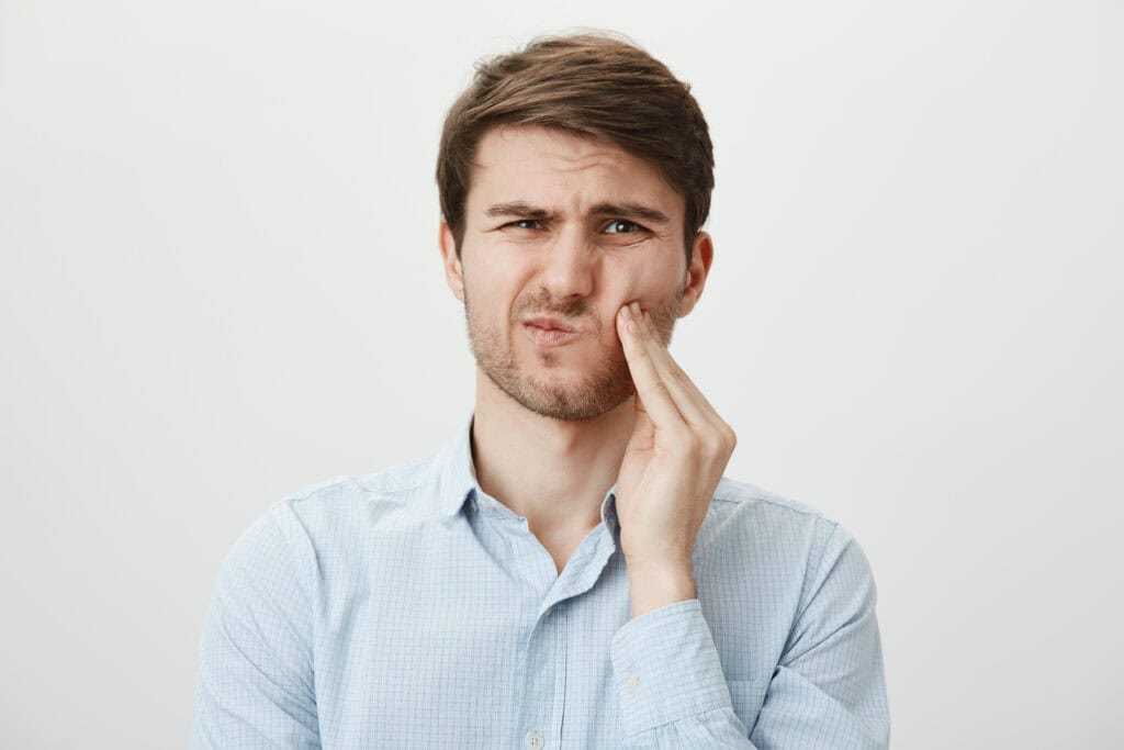 Portrait of handsome young man holding hands over chin, expressing fright and being terrified by his toothache, standing against gray background. Guy can not calm down after witnessing crime scene.