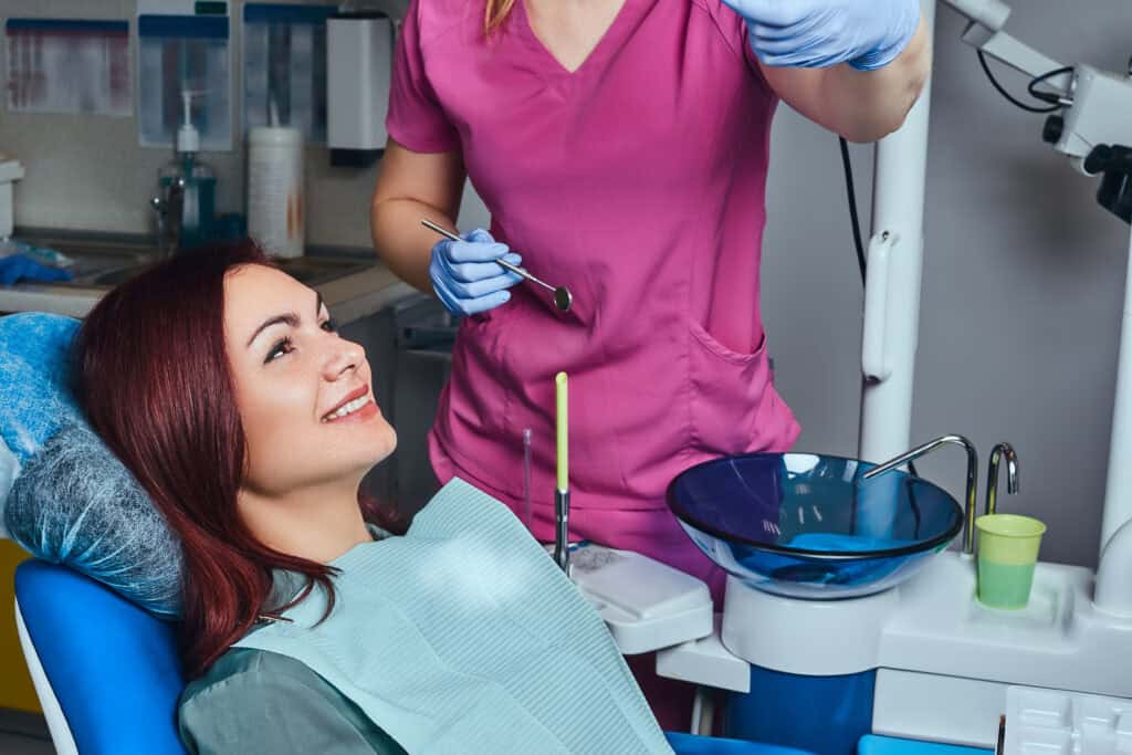 Smiling woman sitting in dental chair during checkup with dentist preparing instruments