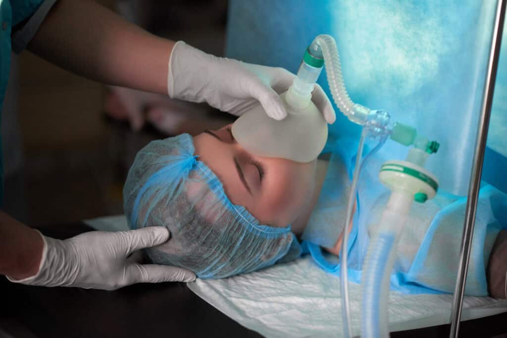 Female patient with a blue surgical cap receiving General Anesthesia via an oxygen mask prior to an oral surgery or complex dental procedure.